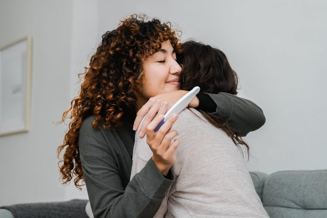 Two women hugging with one holding a positive pregnancy test after using research backed fertility products